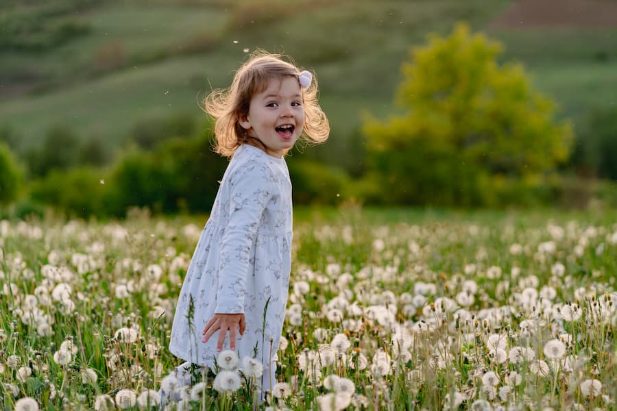 Young child laughing while standing in a field of dandelions, with green hills in the background.