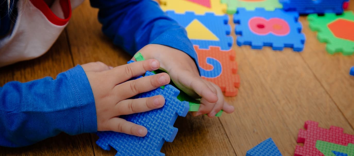 Young child assembling foam puzzle pieces with numbers and shapes on a wooden floor.