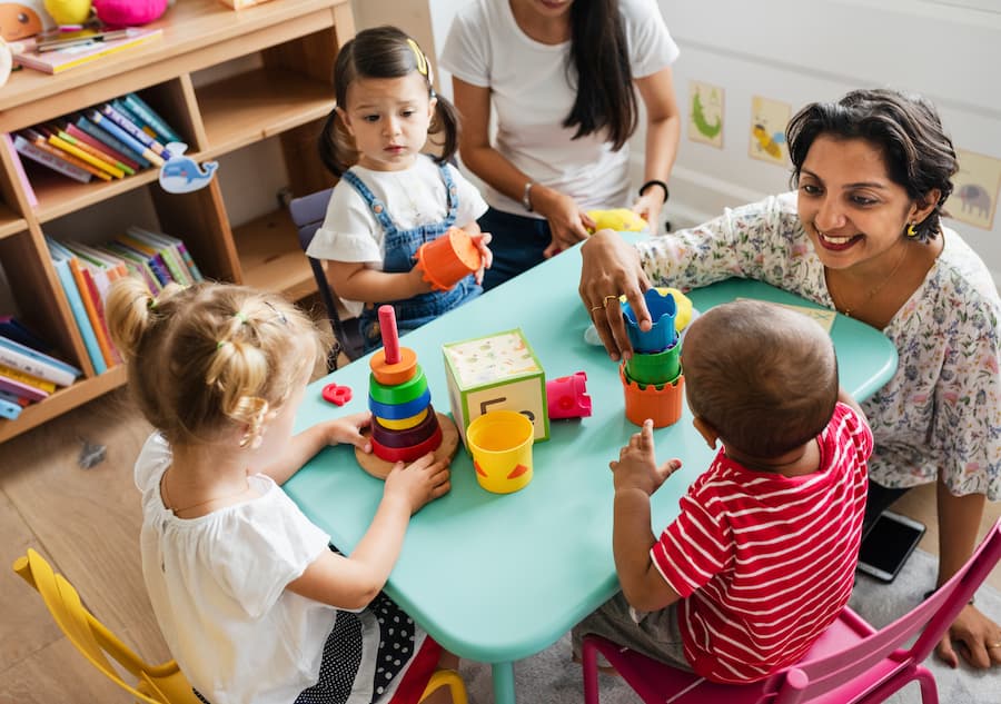 Children playing with stacking toys at a table with two adults in a classroom.