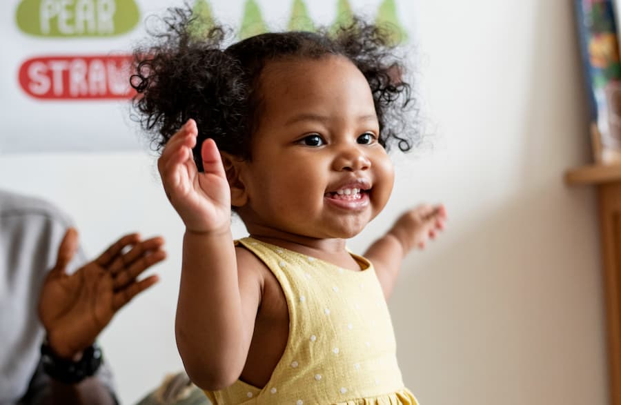 Smiling toddler with arms raised, wearing a yellow polka dot dress, playing indoors.