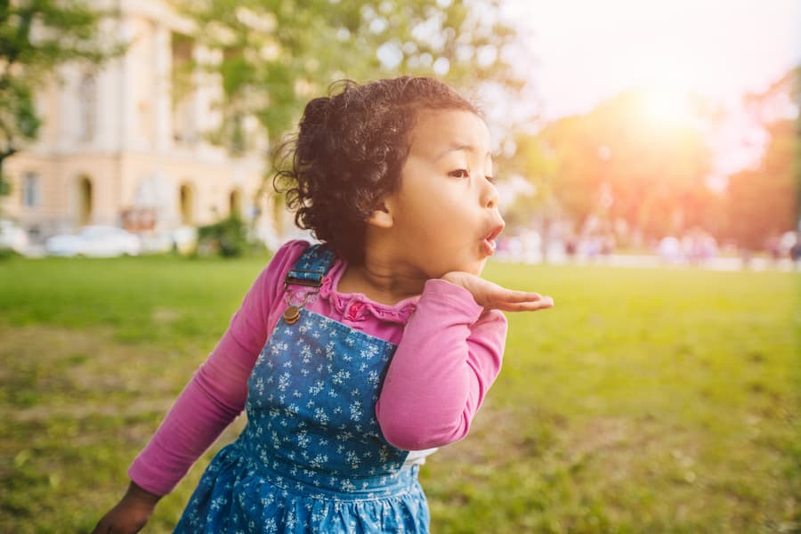 Young child in a pink shirt and denim overalls blowing a kiss outdoors in a sunny park.