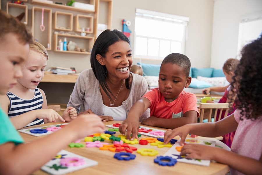 A smiling teacher sits at a table with a diverse group of young children, helping them play and learn with colorful educational toys in a classroom.