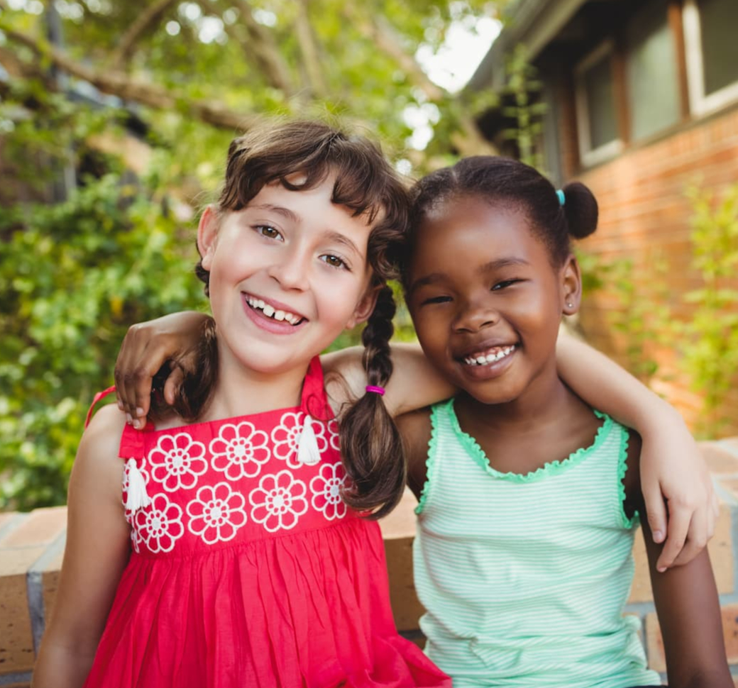Two young girls, smiling with arms wrapped around each others shoulders.